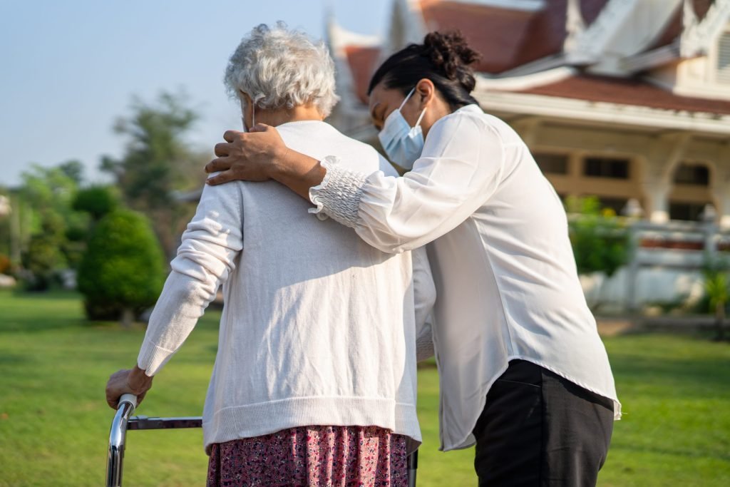 A caregiver walking beside an older woman who is using a walking frame outdoors.