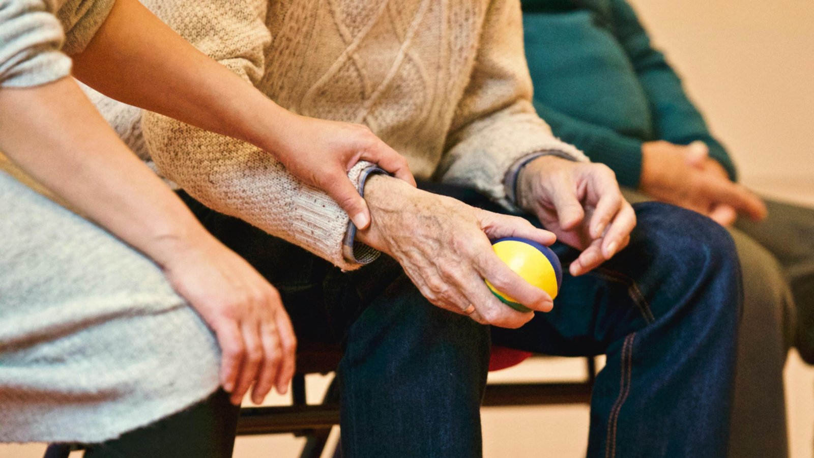A caregiver rests a hand on the arm of an older person who is holding a therapy ball.