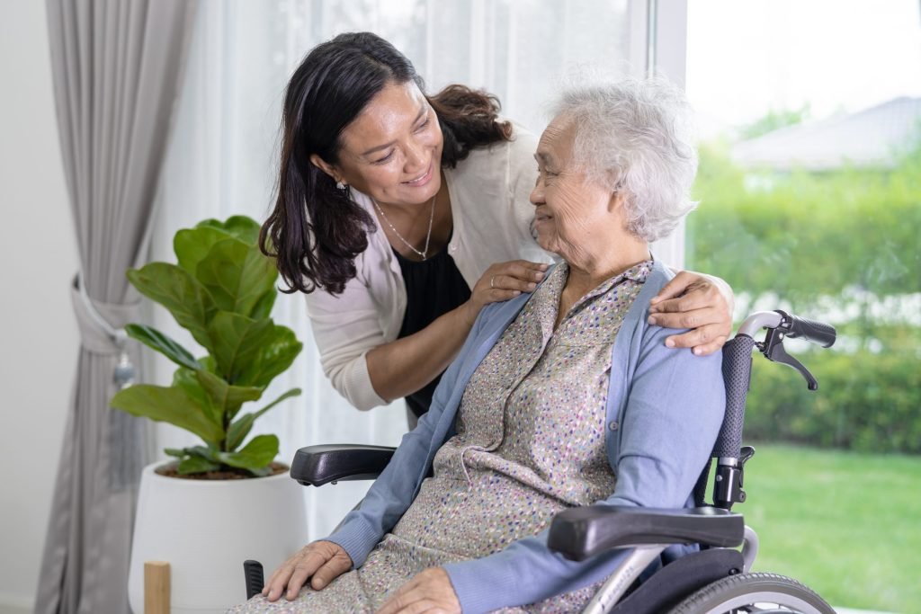 A caregiver assisting an older woman seated in a wheelchair and wearing a face mask.