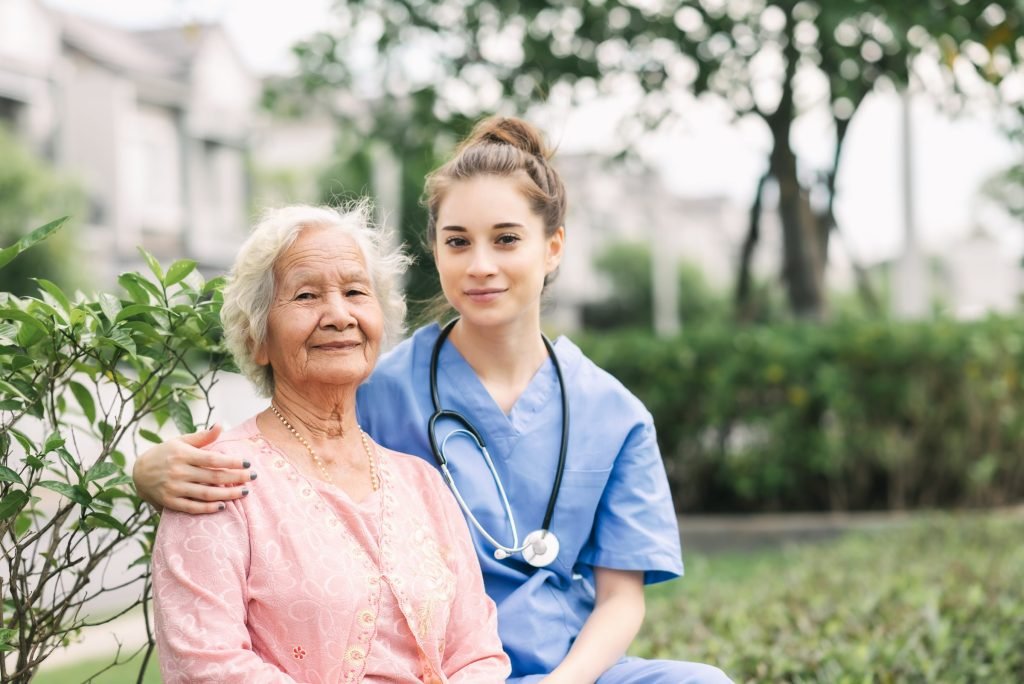 A caregiver with an arm around an older woman, both smiling, in a park.